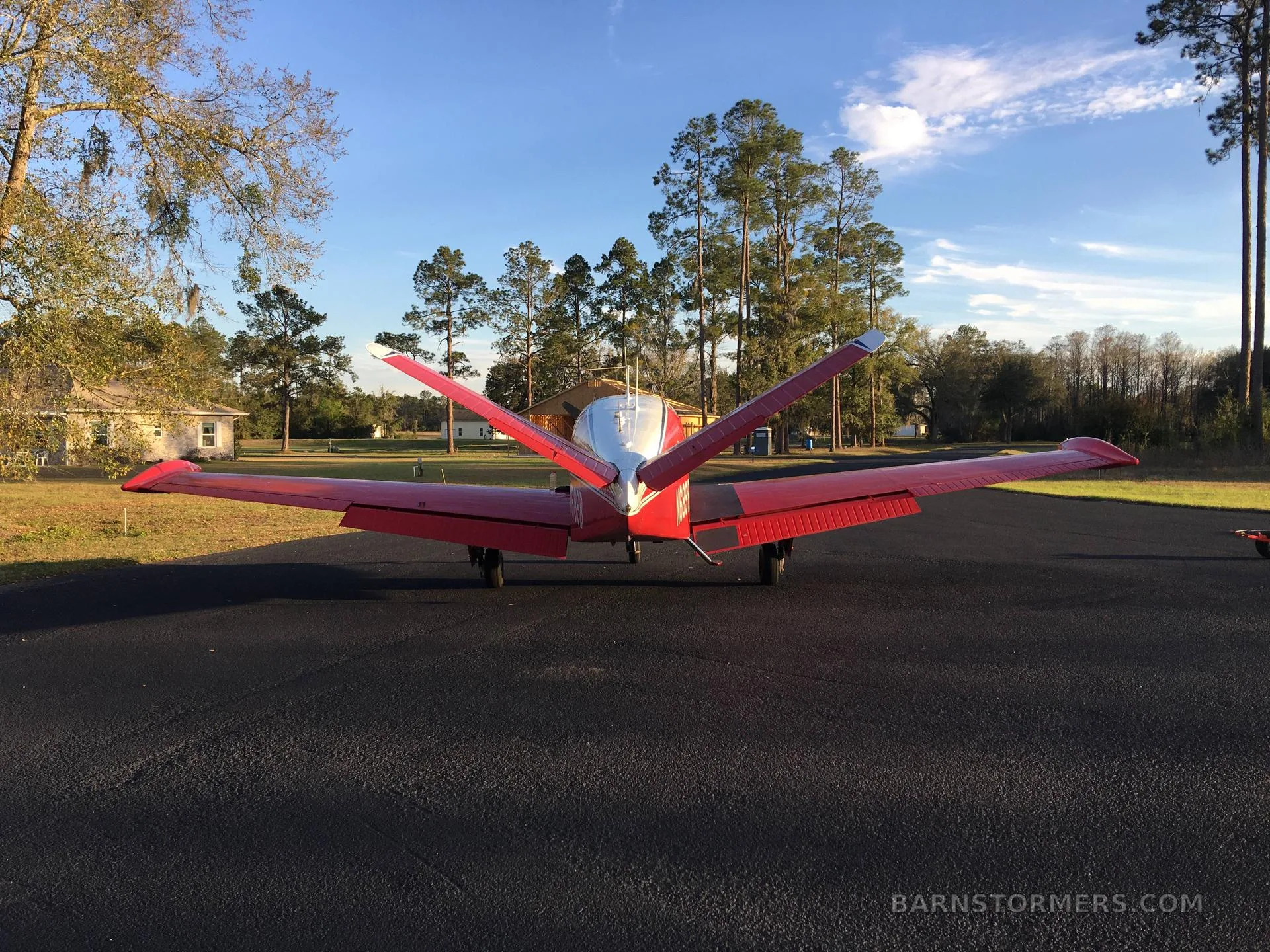 A rear view of a red and white Beechcraft Bonanza parked on an asphalt surface surrounded by pine trees.