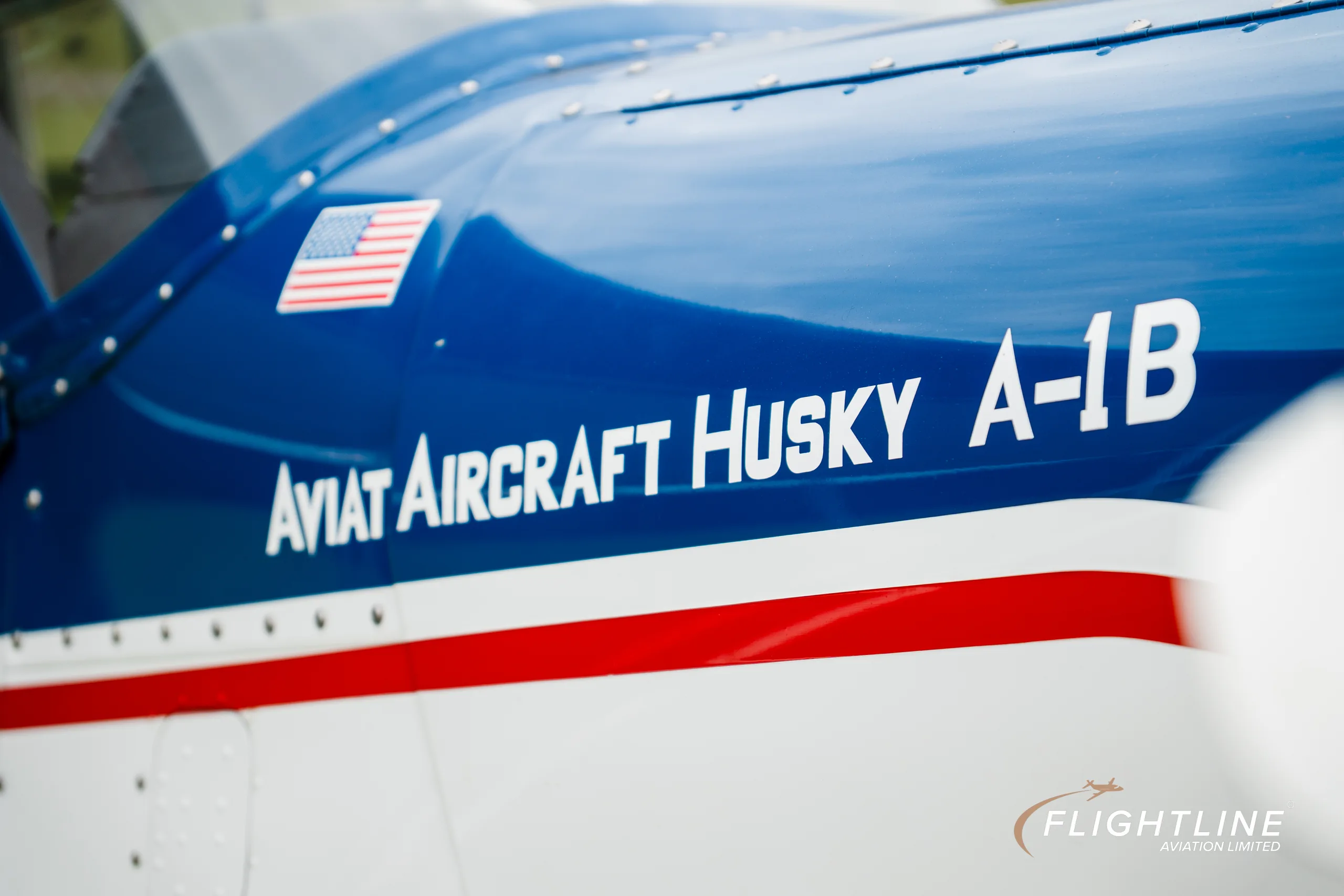 A close-up of the fuselage of the Aviat Aircraft Husky A-1B, showing the aircraft name and a small American flag.