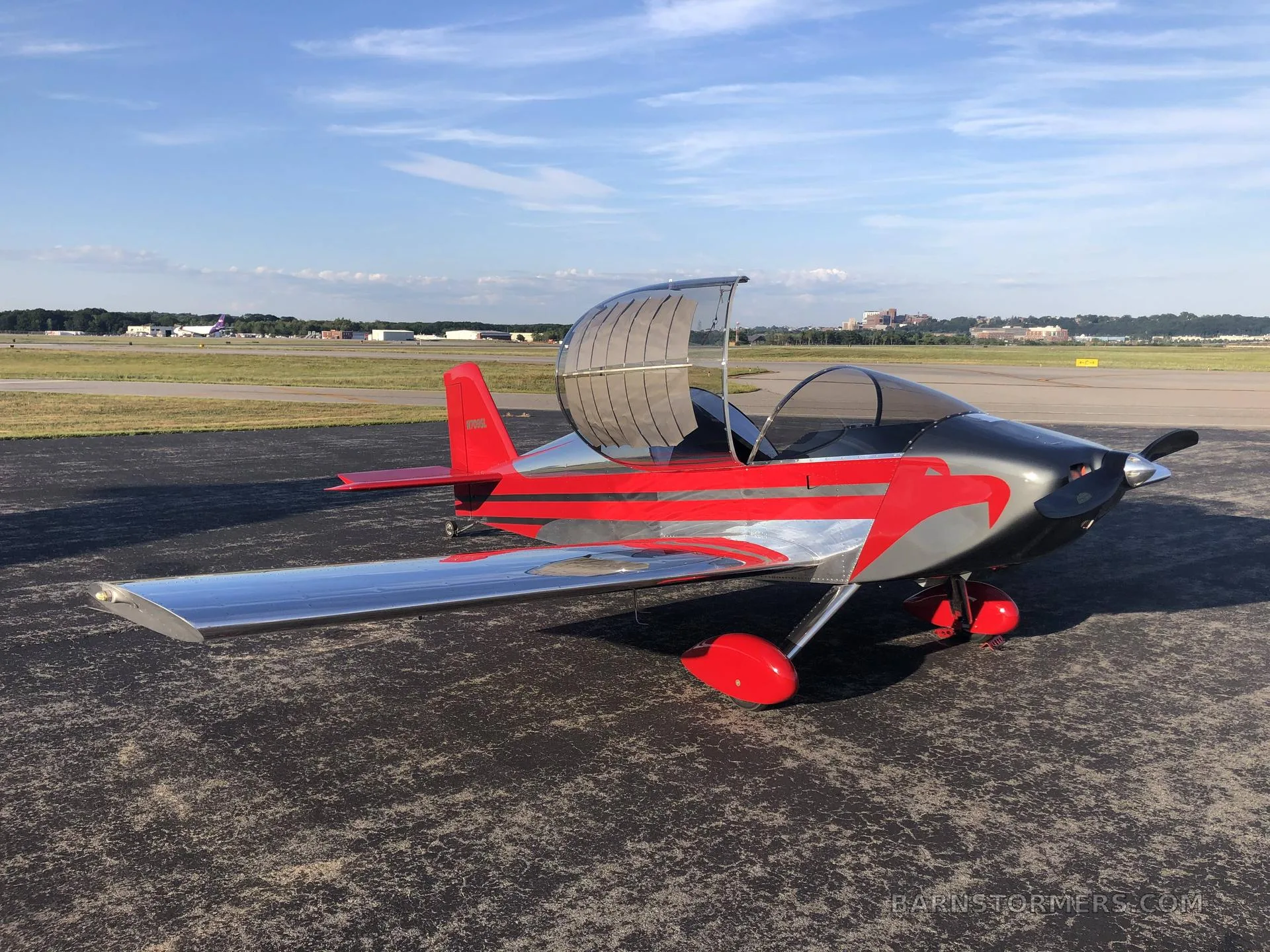 A red and silver experimental aircraft with an open canopy sits on an airport tarmac under a blue sky.