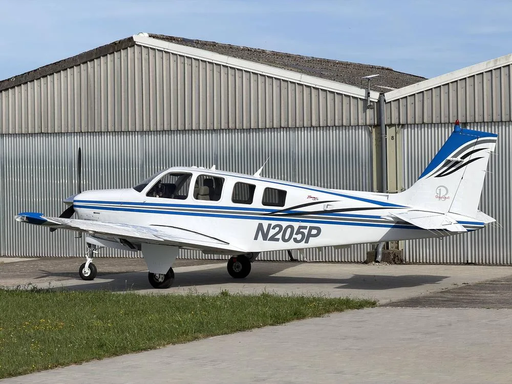 A white and blue Beechcraft Bonanza N205P parked on a paved surface with a hangar in the background.