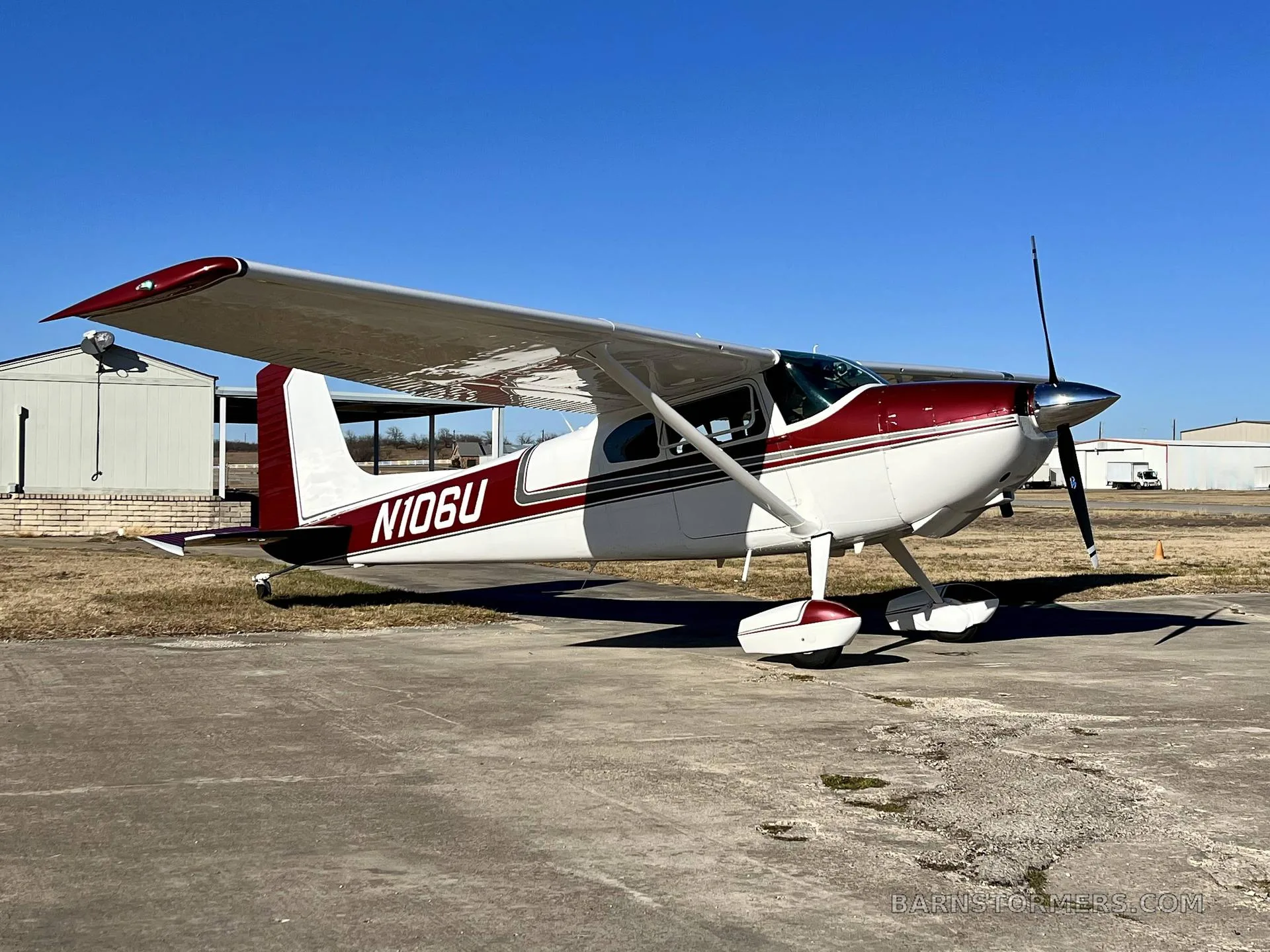 A white and maroon Cessna 182 aircraft (N106U) is parked on a concrete pad next to a hangar under a clear blue sky.