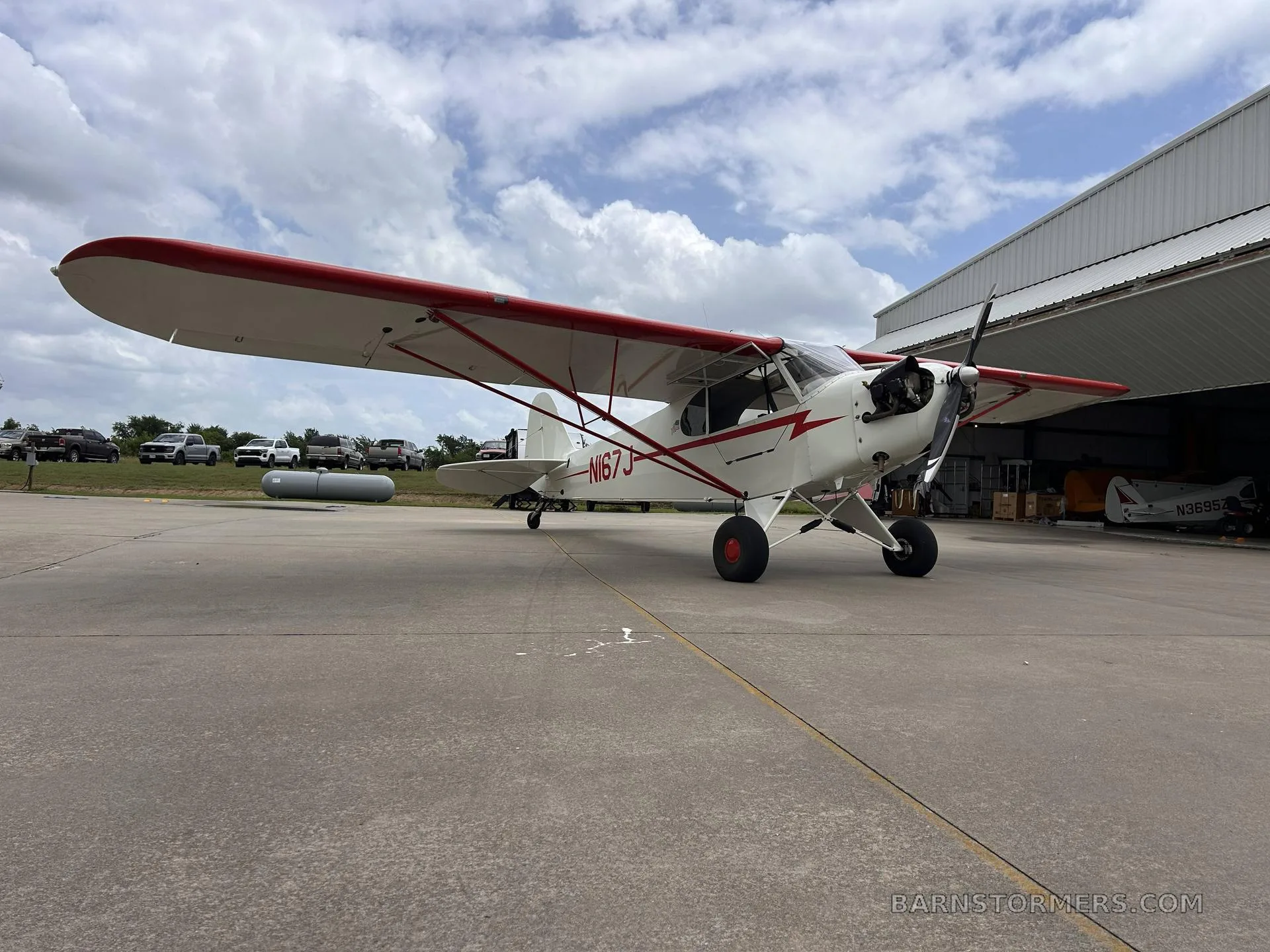 A low-angle view of a white and red single-engine aircraft (N167J) parked on an airfield with hangars and vehicles in the background.