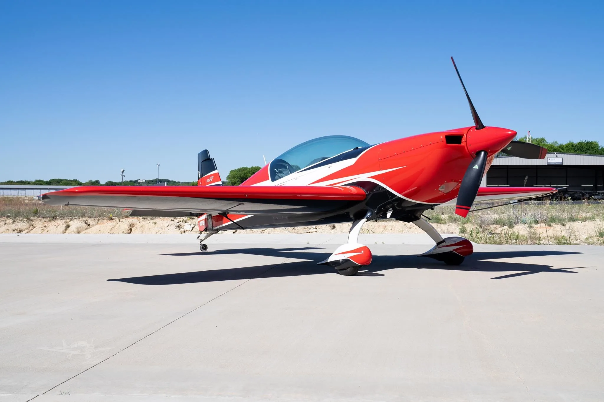 A red, black, and white single-engine propeller aircraft with a bubble canopy is parked on a concrete tarmac under a clear blue sky.