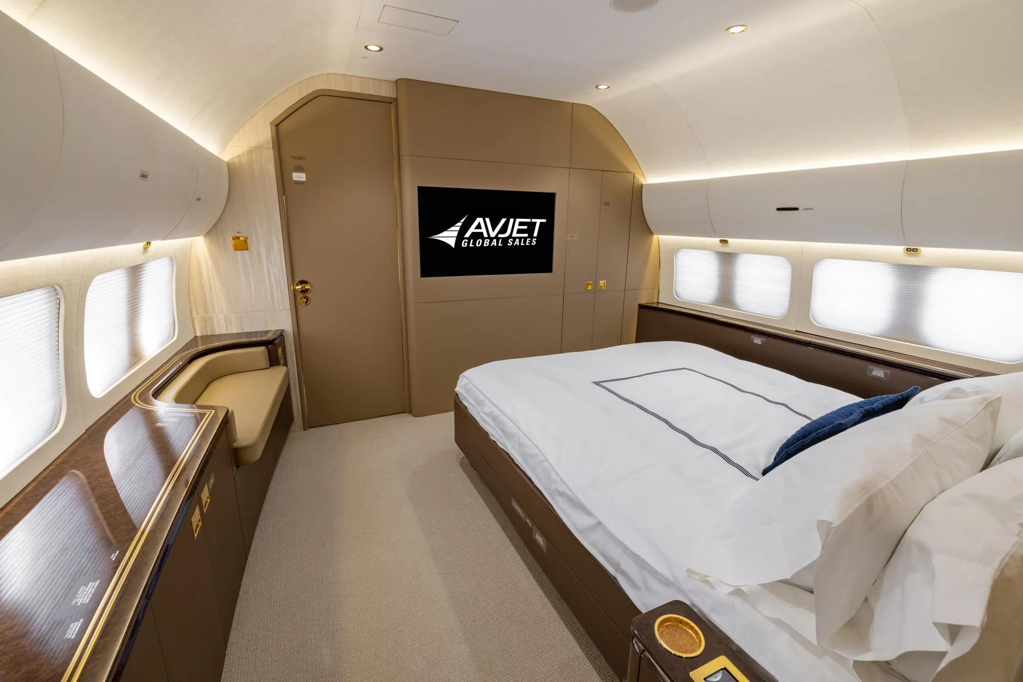 A private jet cabin bedroom featuring a large bed with white linens, a side sofa, and a screen displaying the Avjet logo.