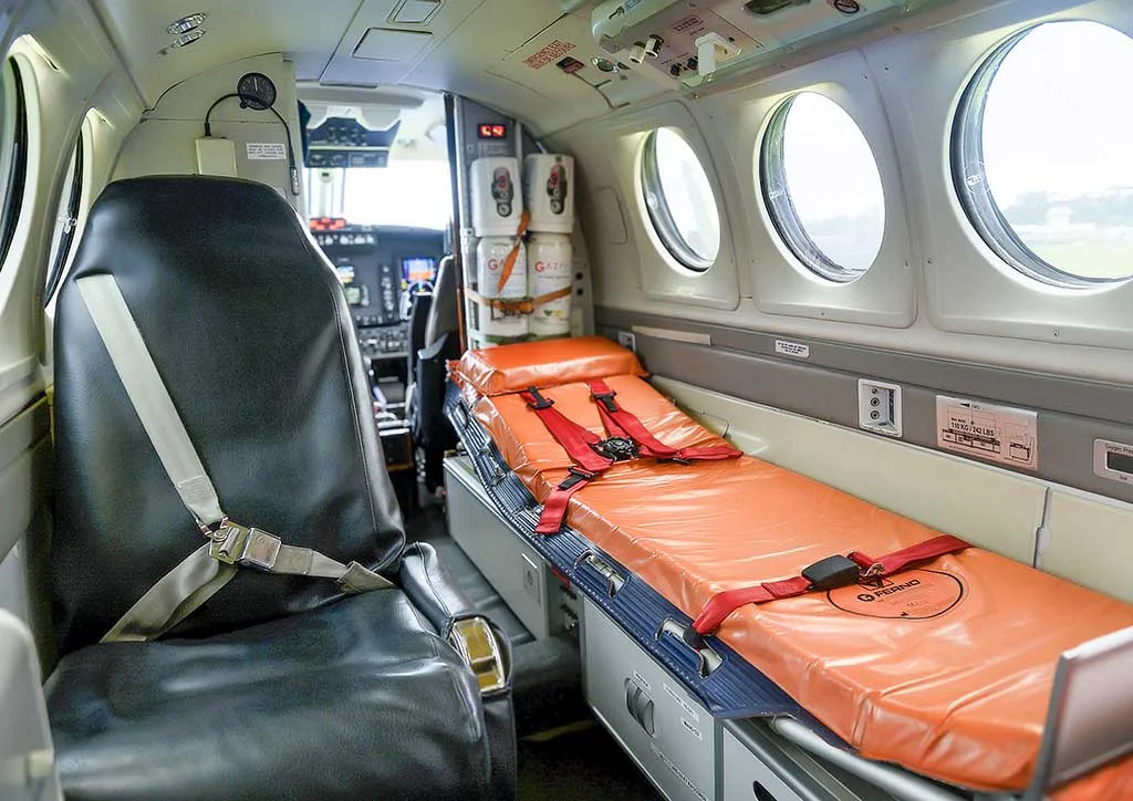 The interior of an air ambulance cabin, featuring an orange medical stretcher and a black passenger seat, with the cockpit visible in the background.