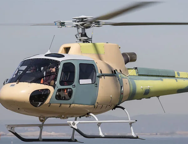 A light utility helicopter with a tan, green, and grey camouflage paint scheme in flight over water.