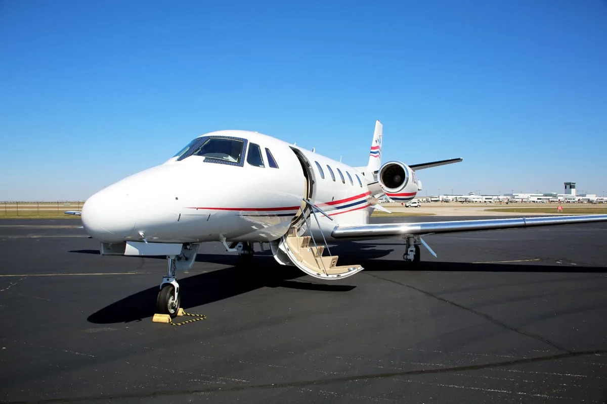 A white business jet with red, white, and blue tail stripes sits on an airport tarmac with its entry door open.