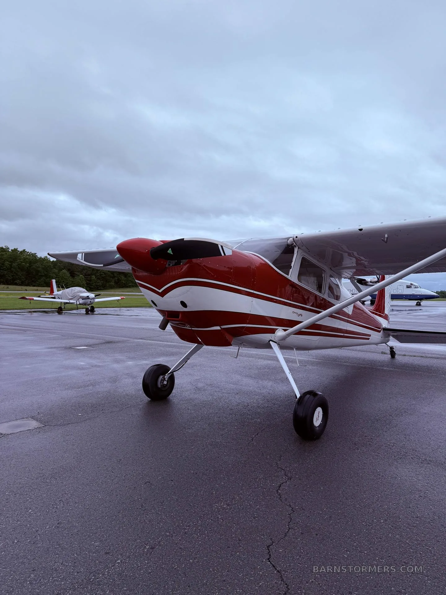 A red and white single-engine propeller aircraft is parked on a wet tarmac, viewed from the front-right.