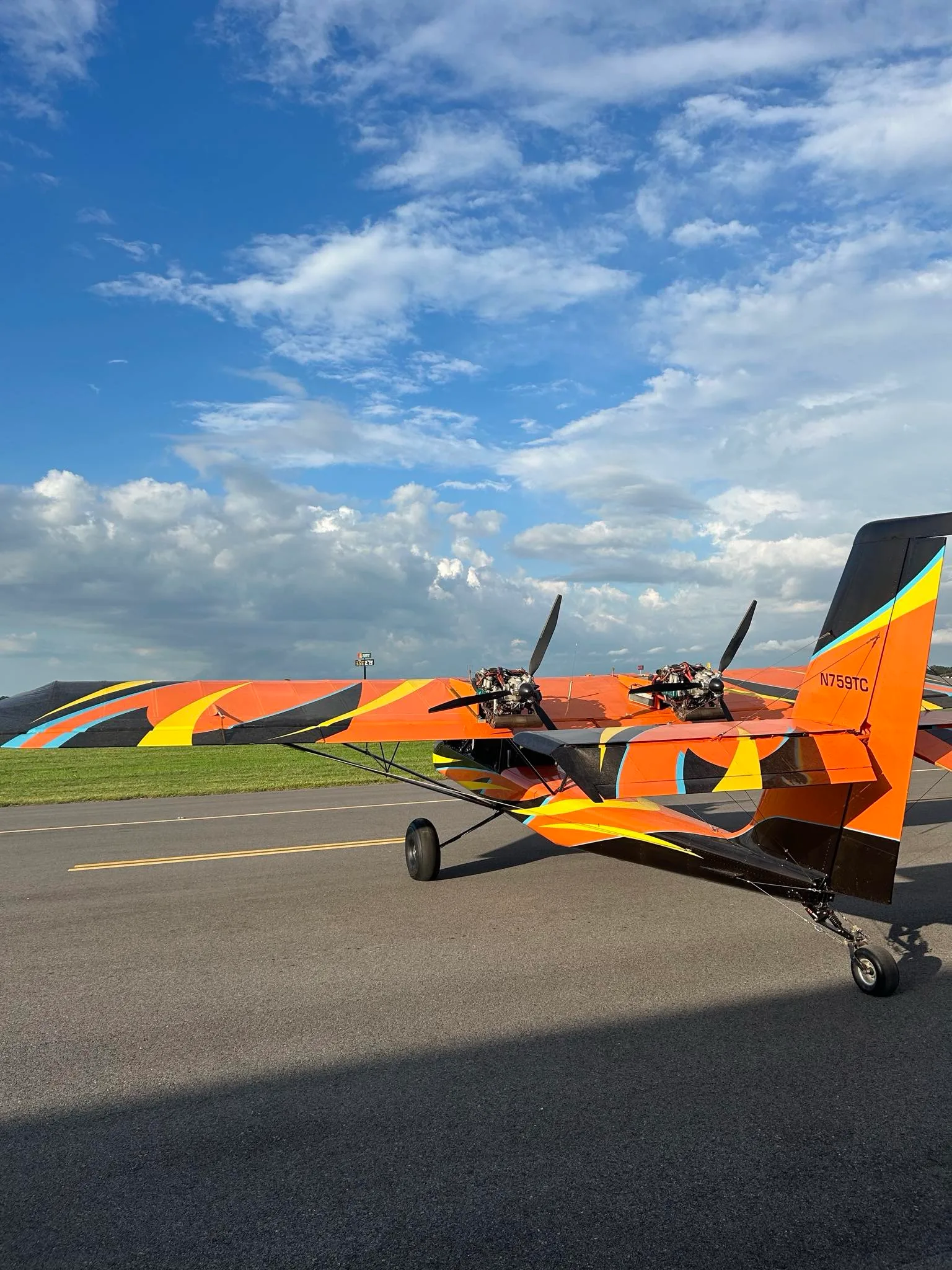 A close-up rear-side view of an orange and black ultralight aircraft, highlighting its twin engines and vibrant paint scheme on the tail and wings.