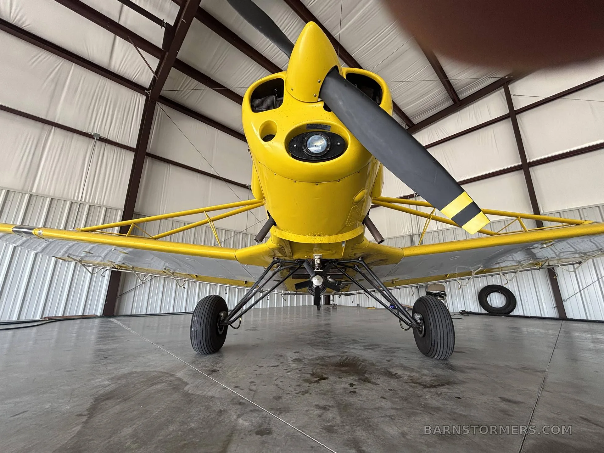 Low-angle front view of a yellow Piper PA-25 Pawnee aircraft with a black propeller, inside a hangar.