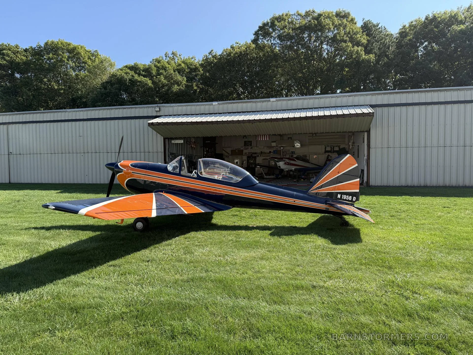 A blue and orange single-engine aircraft with tail number N 1956 D parked on green grass with a hangar open in the background.