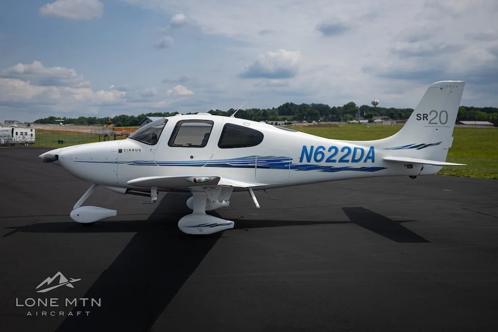 A side view of the white Cirrus SR20 aircraft N622DA with blue accents parked on an asphalt runway with distant buildings.