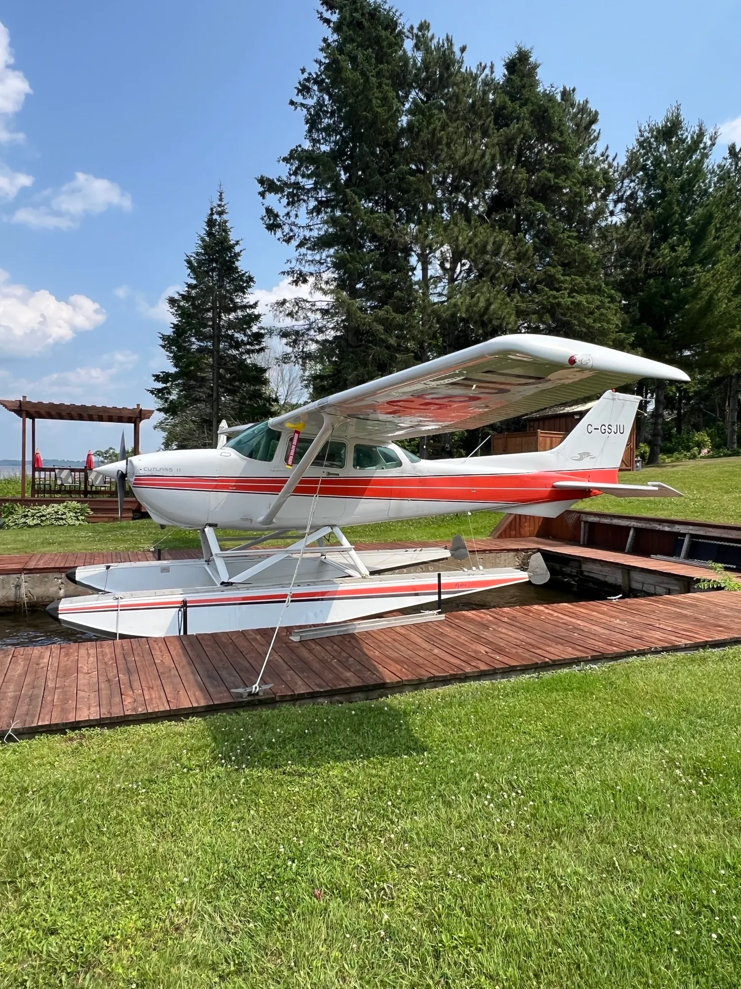 A white and red Cessna Cutlass II floatplane (C-GSJU) is parked on a wooden dock with trees in the background.