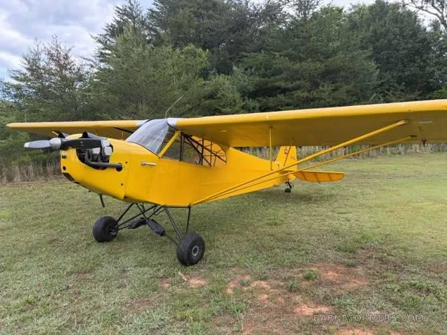 A yellow single-engine taildragger aircraft with an exposed engine, parked on a grassy field with trees in the background.