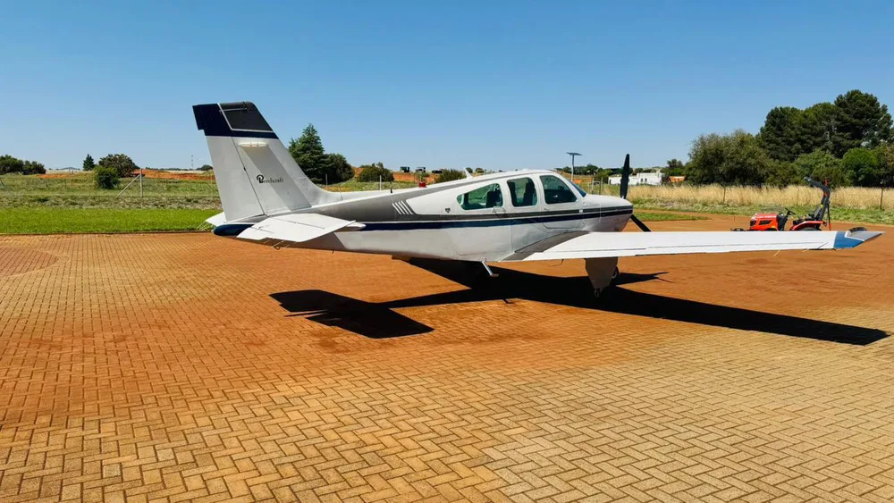A side-rear view of a white and dark grey single-engine propeller aircraft on a brick-paved apron.