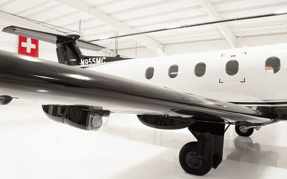 Close-up of the black wing, white fuselage, and landing gear of a Pilatus PC-12 aircraft in a hangar.