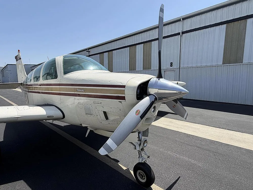 A close-up view of the front left side of a white and gold Beechcraft Bonanza, showing its propeller and nose.