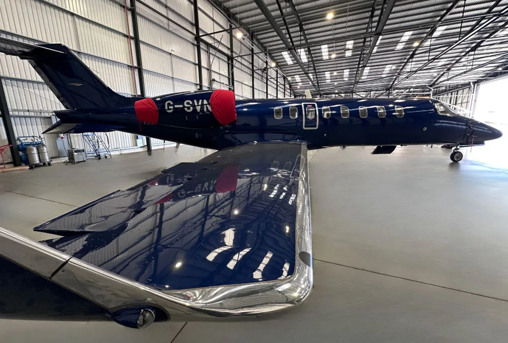 A dark blue private jet with registration G-SVNT and red engine covers parked in a hangar, viewed from the rear-left showing the wing.