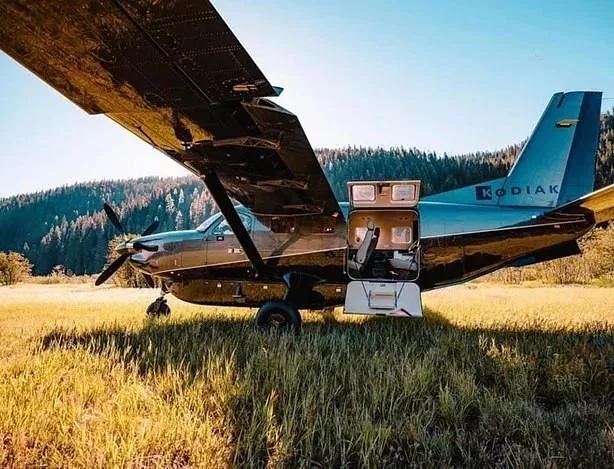 A dark grey and blue Quest Kodiak 100 aircraft with an open cargo door, parked in a sunny grassy field with a forest in the background.