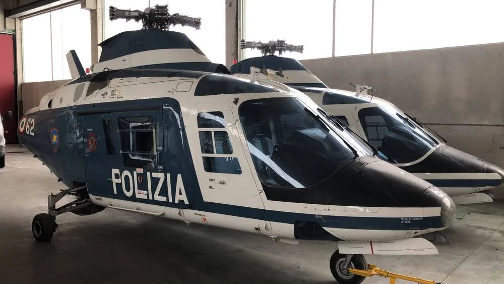 Two blue and white police helicopters, marked "POLIZIA", are parked side-by-side in a hangar.