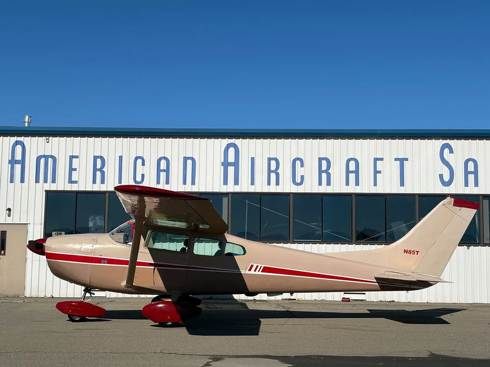 A beige and red Cessna 182 aircraft, registration N85T, parked in front of a hangar with 'AMERICAN AIRCRAFT SA' visible.