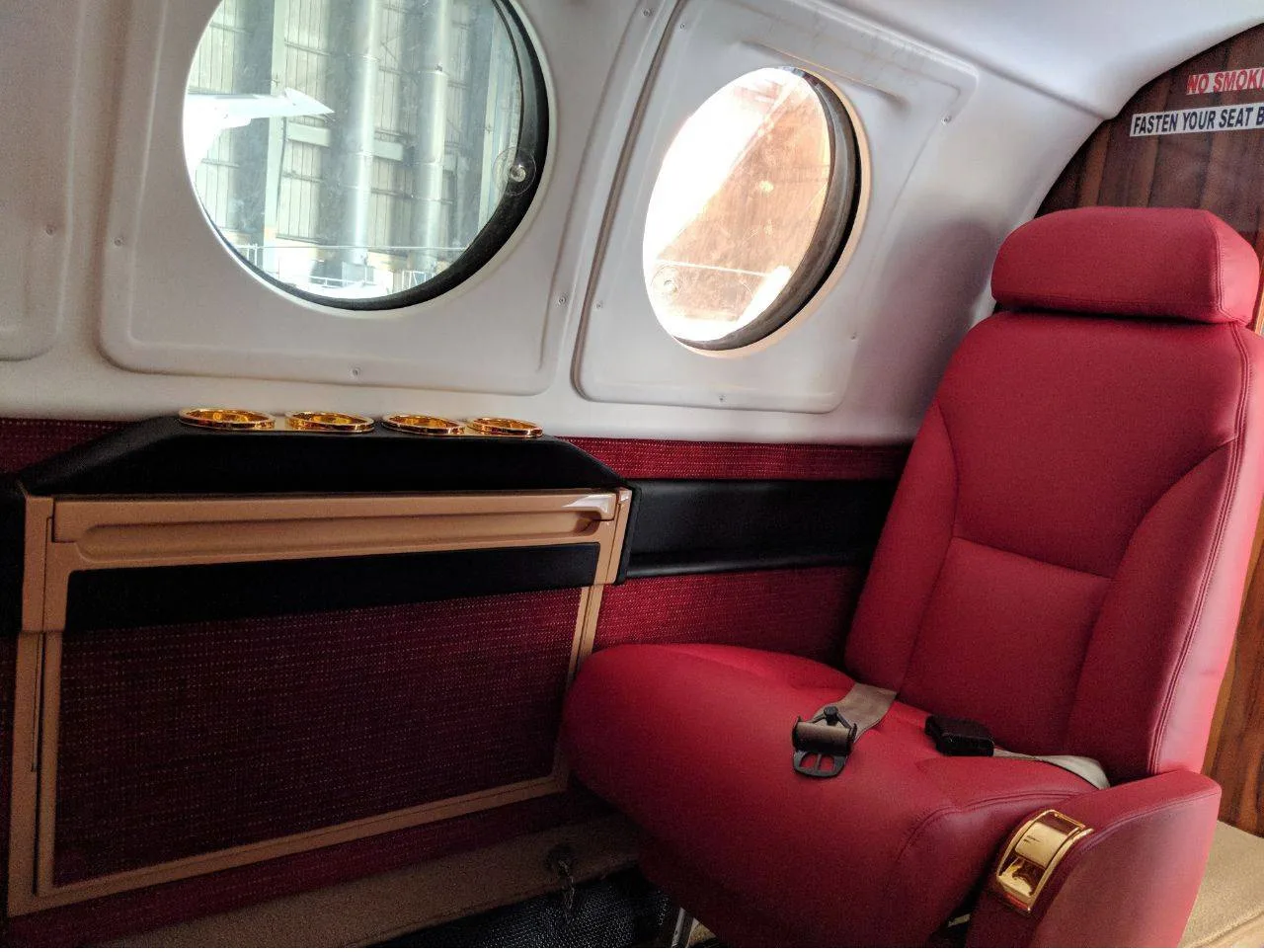 A close-up of a red leather passenger seat next to a side table with cup holders and two oval windows.