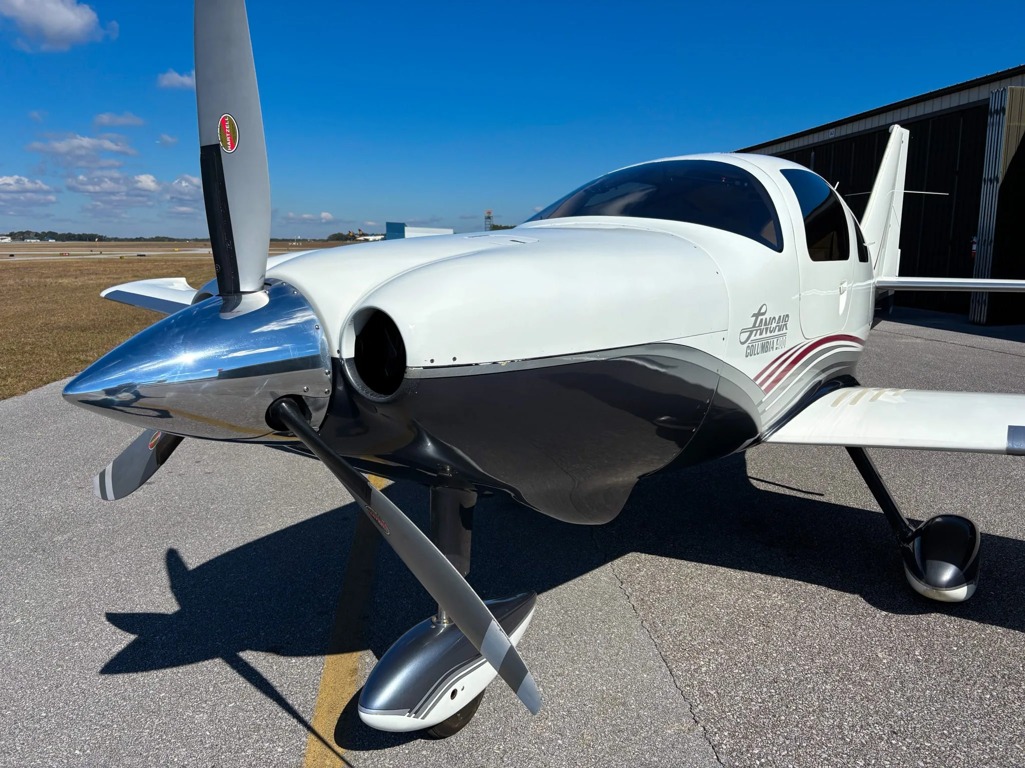 Front-quarter view of a white and grey Lancair Columbia 400 with red stripes on a sunny tarmac.