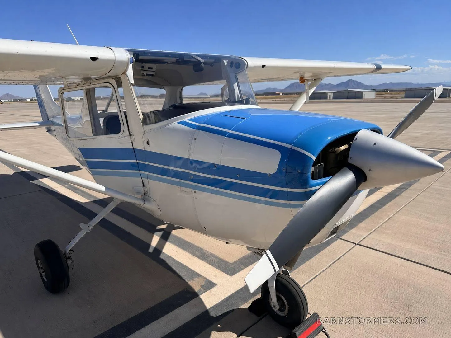 A white and blue single-engine propeller aircraft, possibly a Cessna, parked on an asphalt tarmac with mountains in the background.
