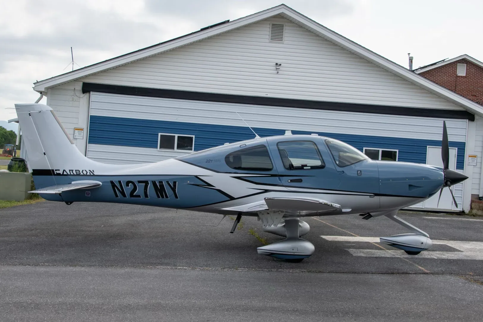 Side view of a blue and white Cirrus SR22T Carbon aircraft, N27MY, parked in front of a hangar.