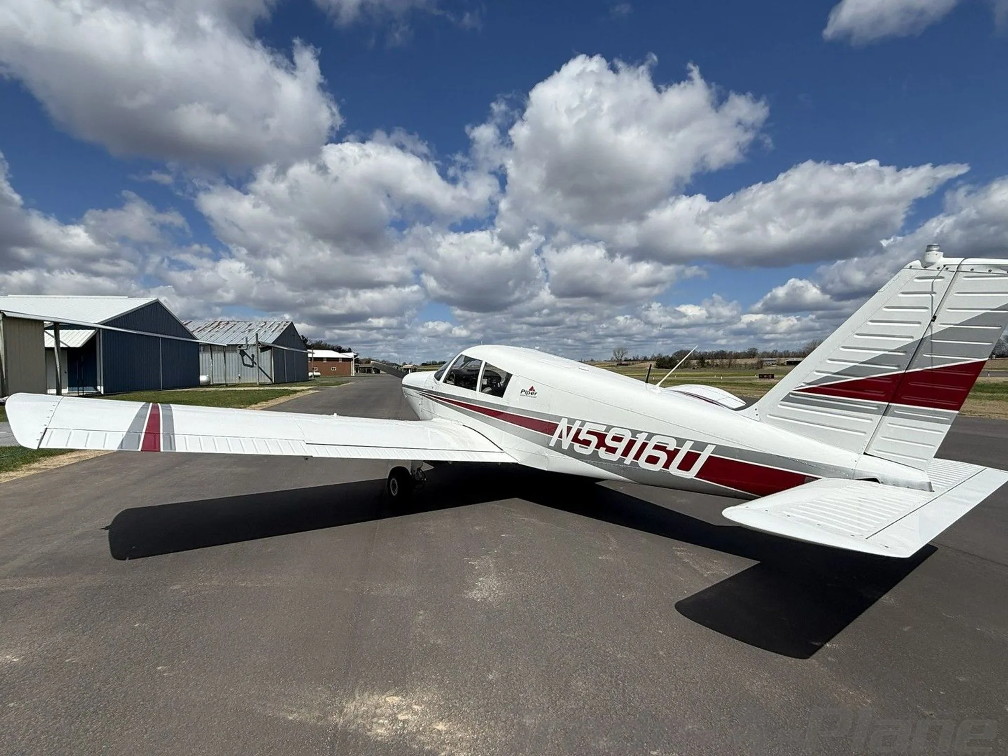 A rear-side view of a white Piper Cherokee aircraft with red and grey stripes on an airfield.