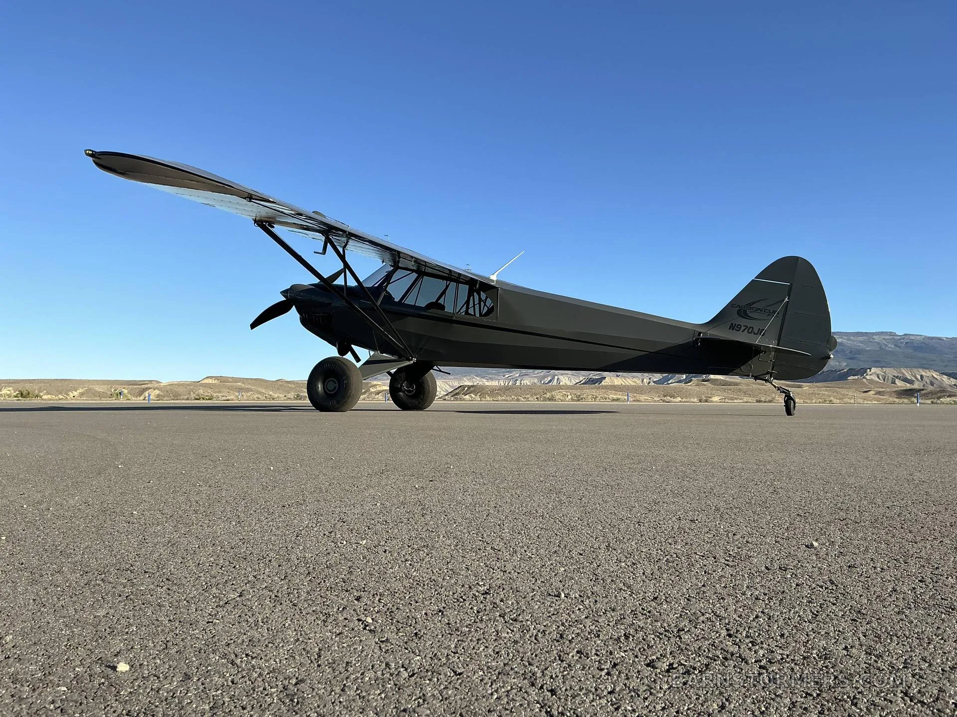 A dark grey Carbon Cub FX3 aircraft with registration N870JH parked on an asphalt runway with mountains in the background.