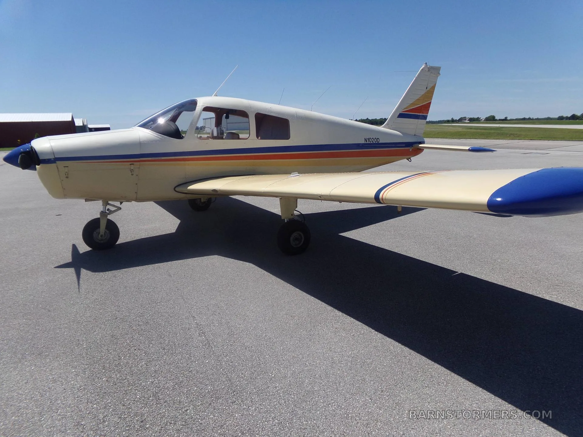A white and beige single-engine Piper aircraft with blue and orange stripes is parked on an asphalt ramp under a clear blue sky.