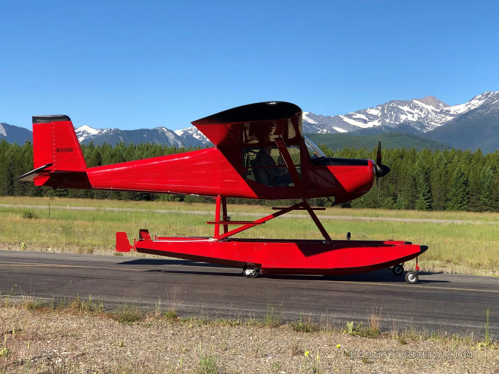 A red floatplane with registration N11286 sits on an asphalt strip with snow-capped mountains in the background.
