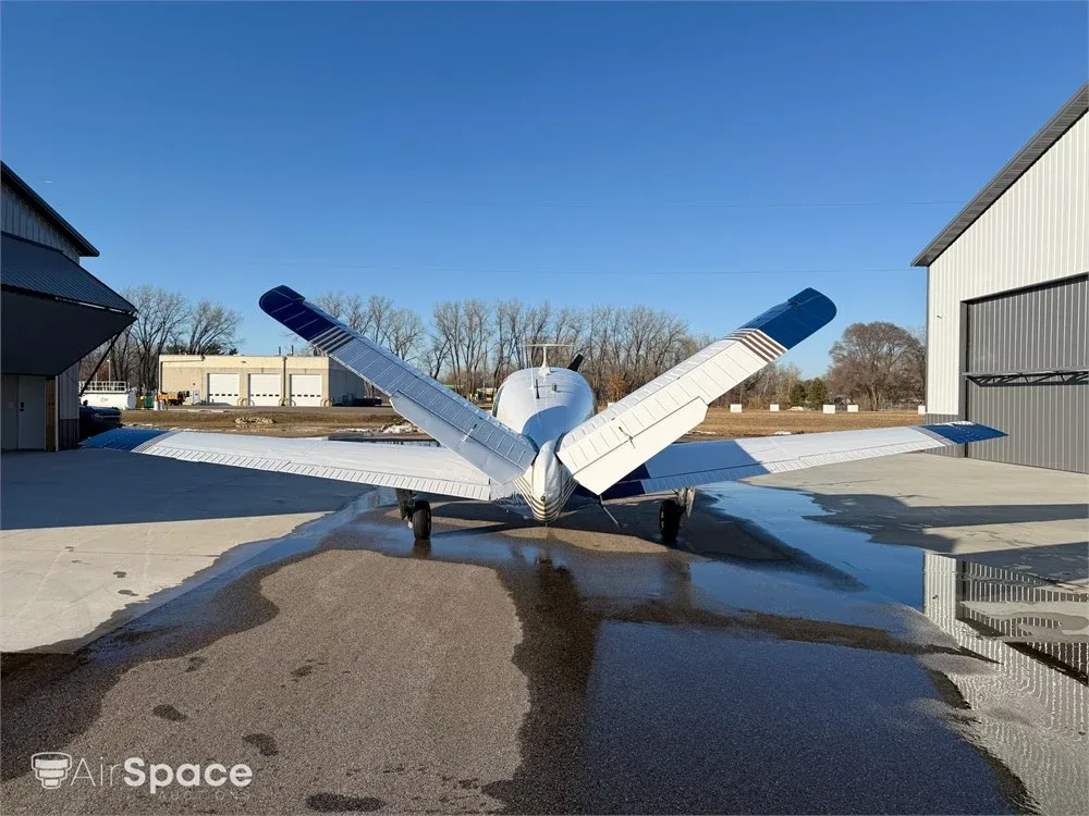 Rear view of a blue and white Beechcraft Bonanza with its V-tail, parked between hangars on a wet surface.