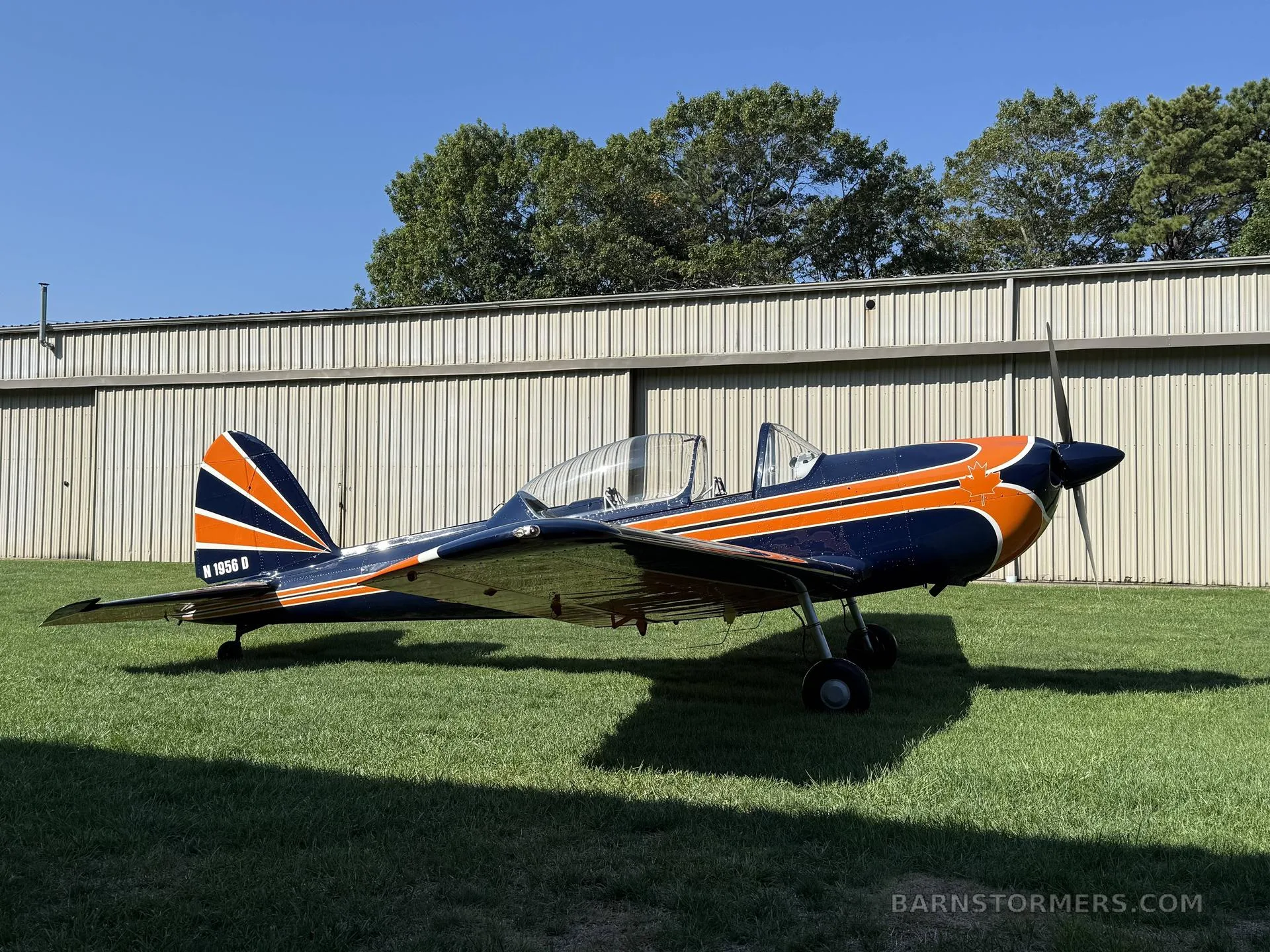 A blue and orange single-engine aircraft with tail number N 1956 D parked on green grass in front of a hangar.