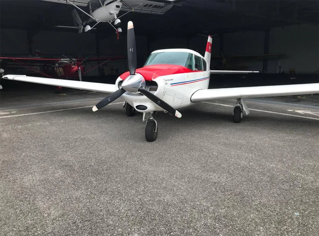 A white single-engine aircraft with a red nose and blue stripes, parked inside a hangar, viewed from the front.