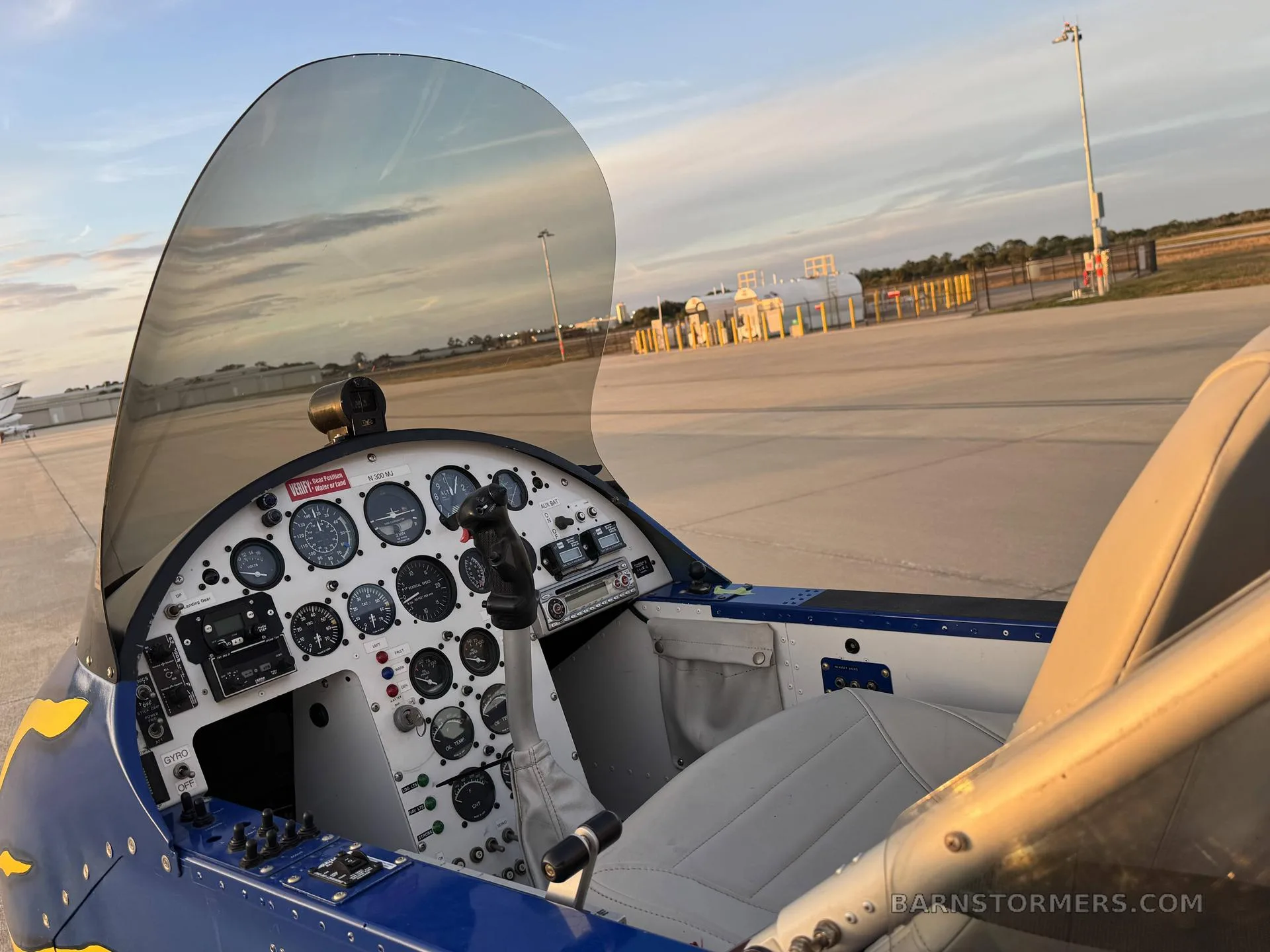 The cockpit of the Air-Cam seaplane, showing the instrument panel, control stick, and pilot's seat with the airfield visible through the windshield.
