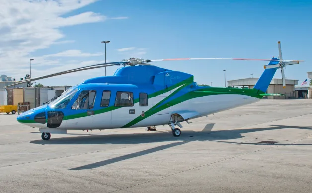 A blue, green, and white helicopter, likely an AgustaWestland AW139, is parked on an airfield tarmac under a partly cloudy sky.