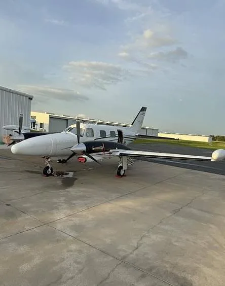 A three-quarter front view of a white and black twin-engine propeller aircraft with red propeller covers, parked on a tarmac next to a hangar.