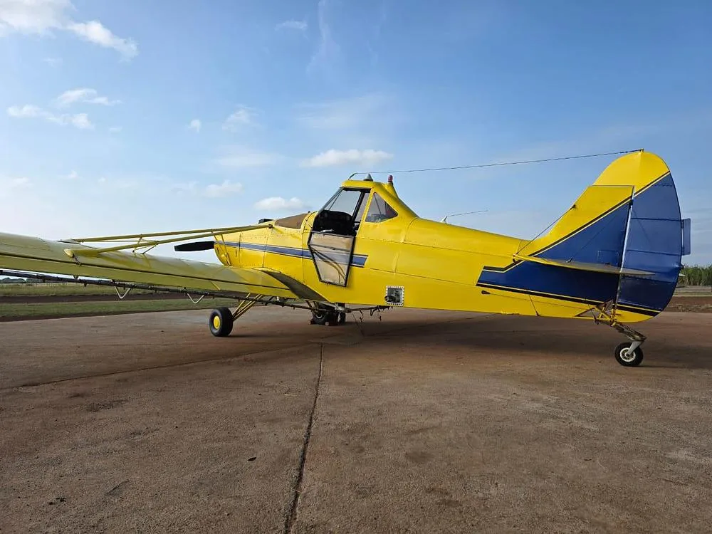 A yellow agricultural aircraft with blue stripes and an open door is parked on a concrete pad.