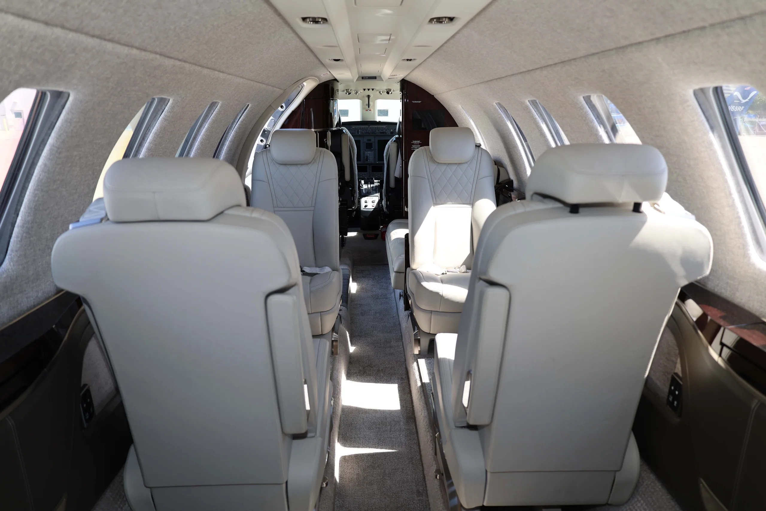 A view down the aisle of a private jet cabin, showing multiple light-colored leather seats and the cockpit beyond.