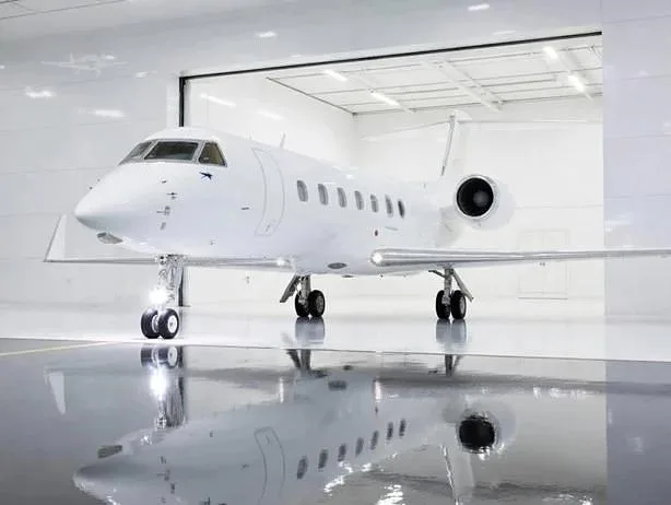 A sleek white business jet with rear-mounted engines is parked inside a brightly lit hangar with a reflective floor.