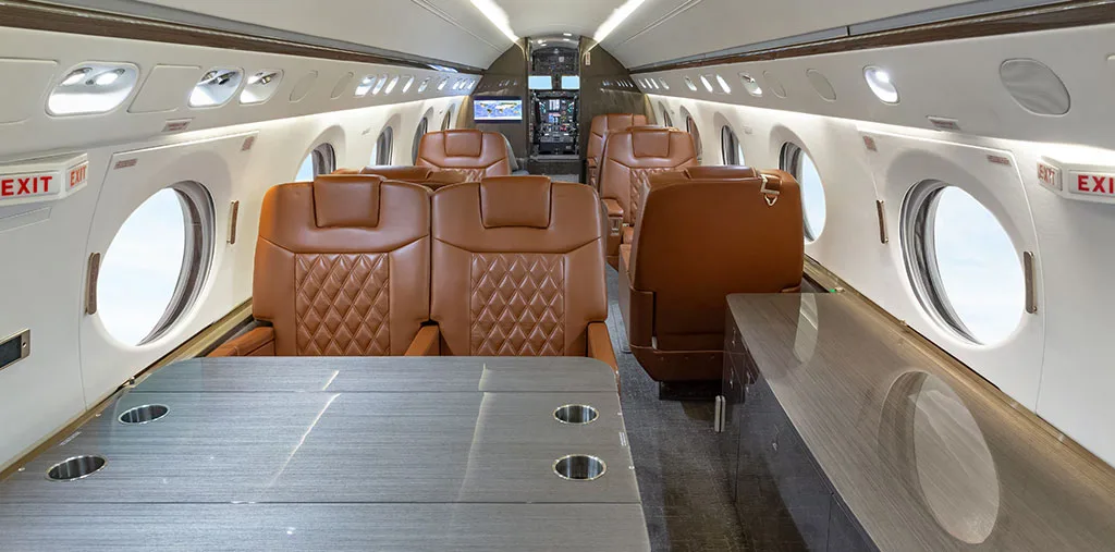 The spacious interior of a private jet cabin with brown leather seating and a table, looking towards the cockpit.