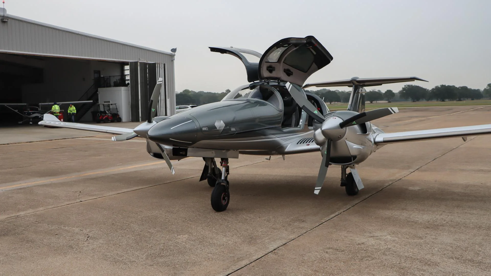 A front-side view of the gray and silver Diamond DA62 aircraft with its gull-wing doors open, parked on an airport tarmac.