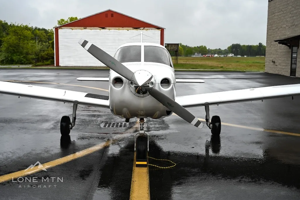 Front view of a white single-engine propeller aircraft with a silver nose, parked on a wet asphalt tarmac in front of a red and white hangar.