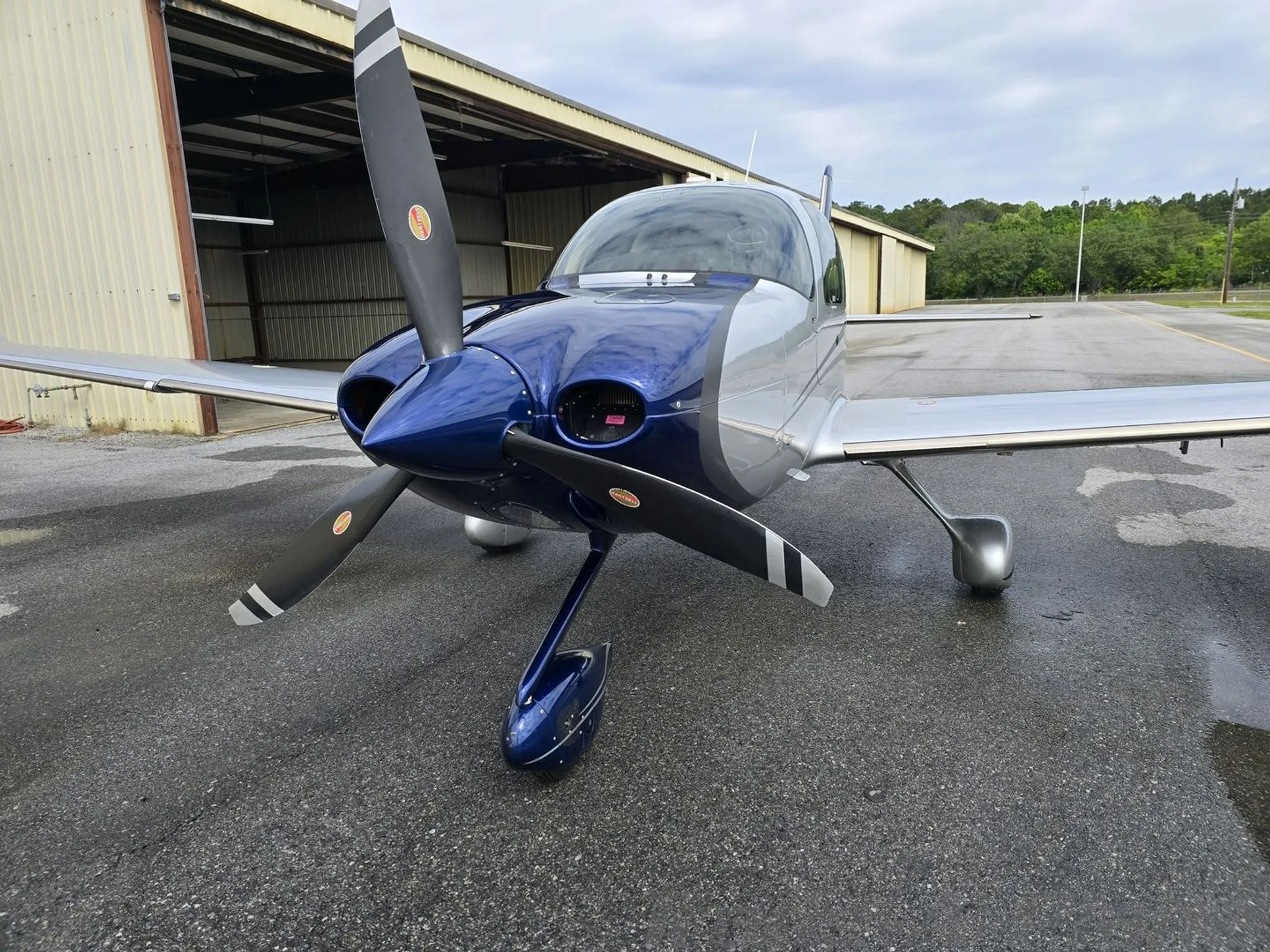 Front view of a blue and silver Cirrus SR22T aircraft with its propeller, parked on a wet tarmac.