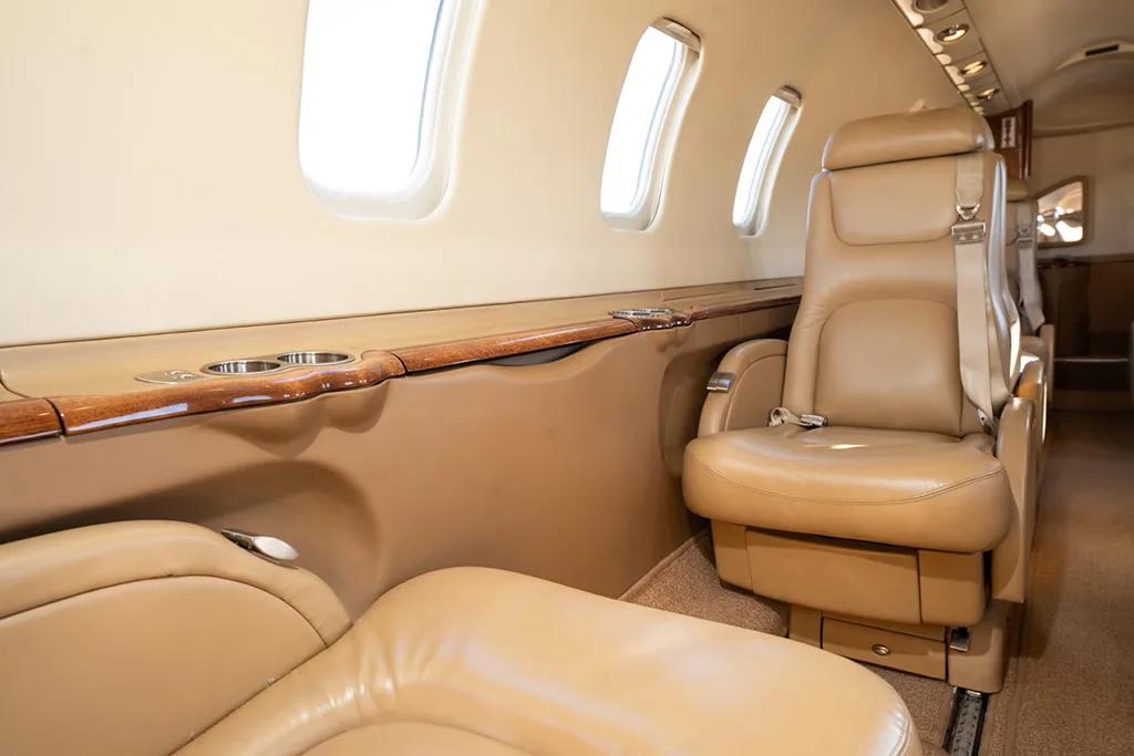 Close-up of a beige leather cabin seat next to a window and a wooden side panel with cup holders.