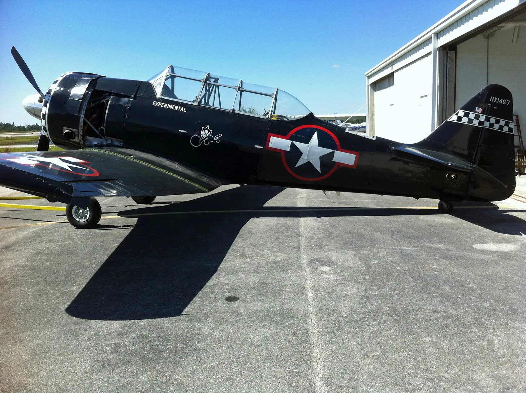 A black warbird aircraft with a checkered nose and US star insignia, registration NX1467, parked on an airfield.