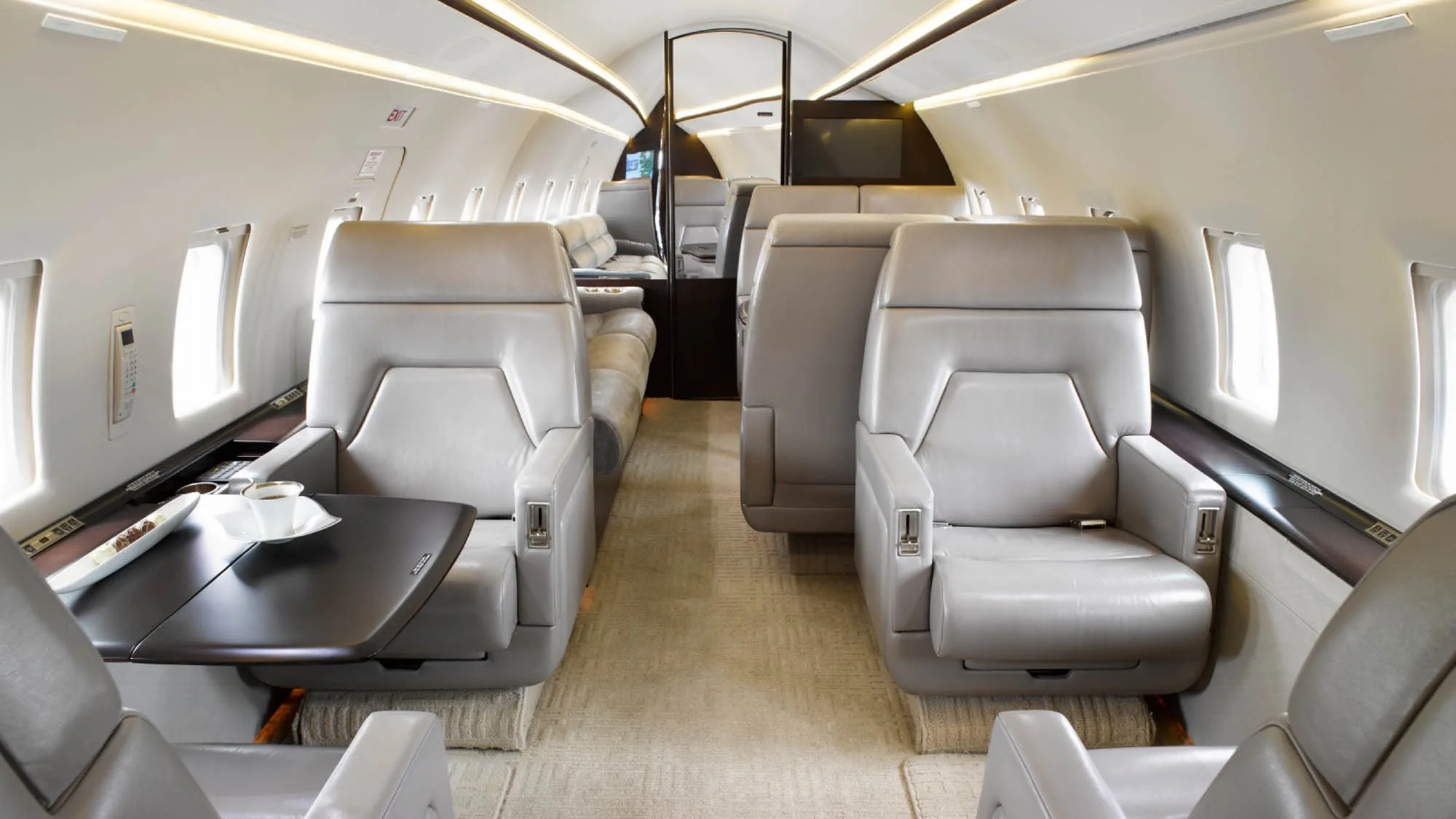 The luxurious cabin interior of a business jet, featuring light grey leather seats and a dark wood table.