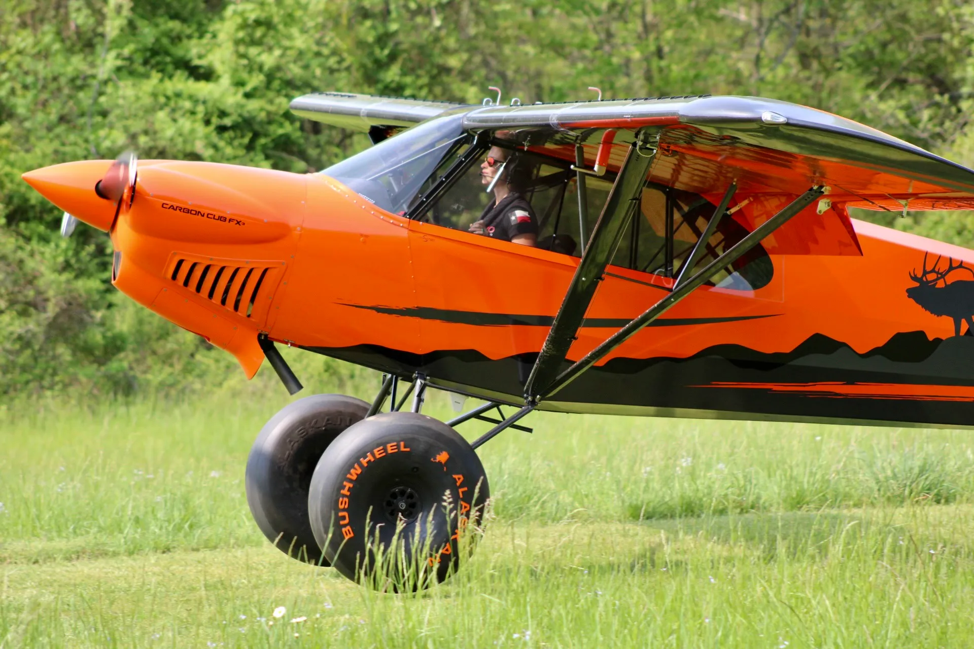 A pilot in an orange and black Carbon Cub FX-3 aircraft with large bush wheels, on a grassy field.
